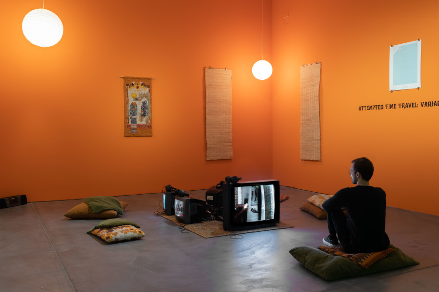Renée Green, Übertragen/Transfer, 1997, Installation. Mixed media: 16 color photographs, six b/w photographs, three lithographs, vinyl type on wall (Attempted Time Travel Variable Sites Continuing Circuit “Complex Fate”), five videos (Partially Buried, 1996; Conversation with Janice and Lonnie “Lumumba” Turner, 1996; Übertragen/Transfer (Samir Alschausky, Henrietta Schroeter, Simin Farkhondeh), 1996; Super 8 Footage, 1996; Underground, 1976, a film by Emile de Antonio, Mary Lampson, and Haskell Wexler), sound, one hanging appliqué, two paper ball lamps, two window roller shutters, sisal carpet, pillows, wall treatment. Courtesy Museum Ludwig, Köln / Ankauf mit Unterstützung der Ludwig Stiftung 2001. Photo: Flavio Karrer.