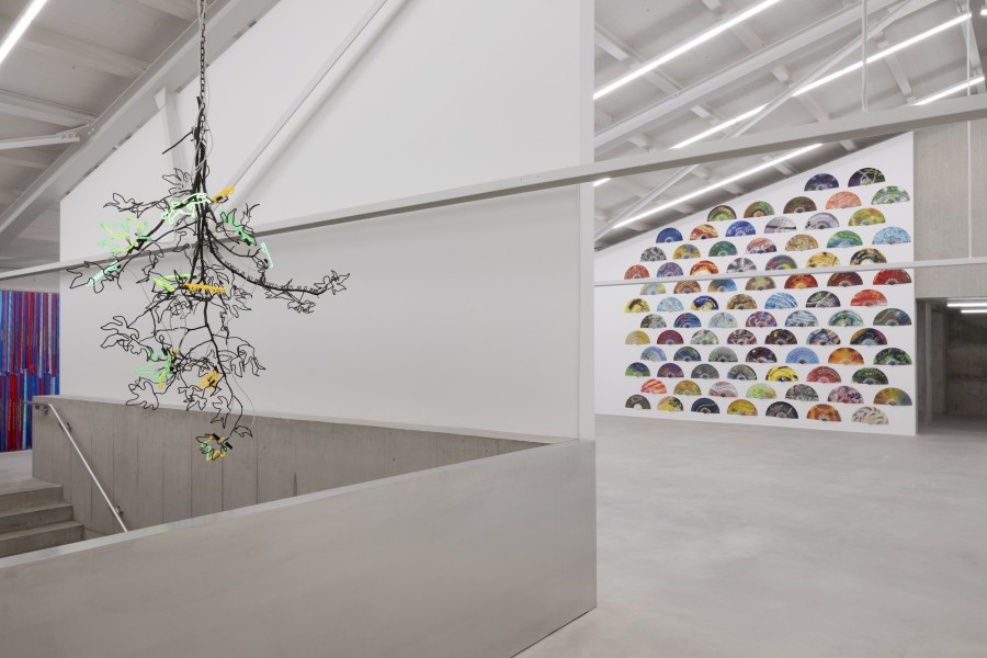 Andrea Bowers, Chandeliers of Interconnectedness (The world is so beautiful even as it burns; Quote used by permission of Terry Tempest Williams), 2022. Steel, neon lights, 185 x 120 x 100 cm. “The world is so beautiful even as it burns” copyright ©2019 by Terry Tempest Williams, from the book EROSION: ESSAYS OF UNDOING. Courtesy of the artist and kaufmann repetto Milan / New York, © 2024, ProLitteris, Zurich; Andrea Bowers, Feminist Fans 2022. Spraypainted fabric fans, 83 fans. Site specific installation, variable dimensions each: 34,2 x 63,5 cm, edition of 5 + 2 AP. Courtesy of the artist and Fondazione Furla and kaufmann repetto Milan / New York, © 2024, ProLitteris, Zurich. Ausstellungsansicht / Installation view Kunsthaus Baselland 2024. Foto / Photo: Gina Folly